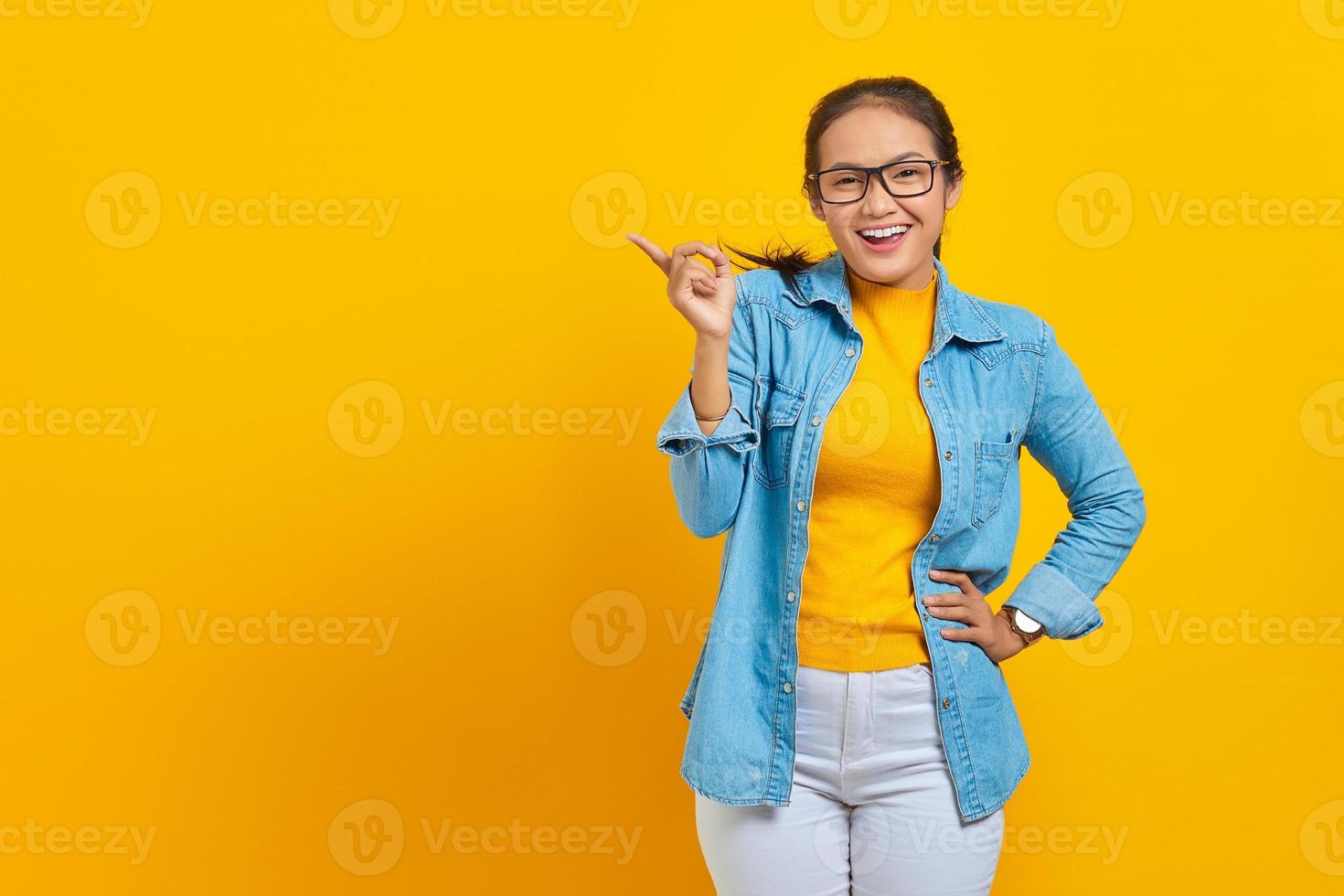 retrato de estudante asiática jovem alegre em roupas jeans, apontando o dedo no espaço da cópia, mostrando produtos publicitários isolados em fundo amarelo. educação no conceito de universidade universitária foto