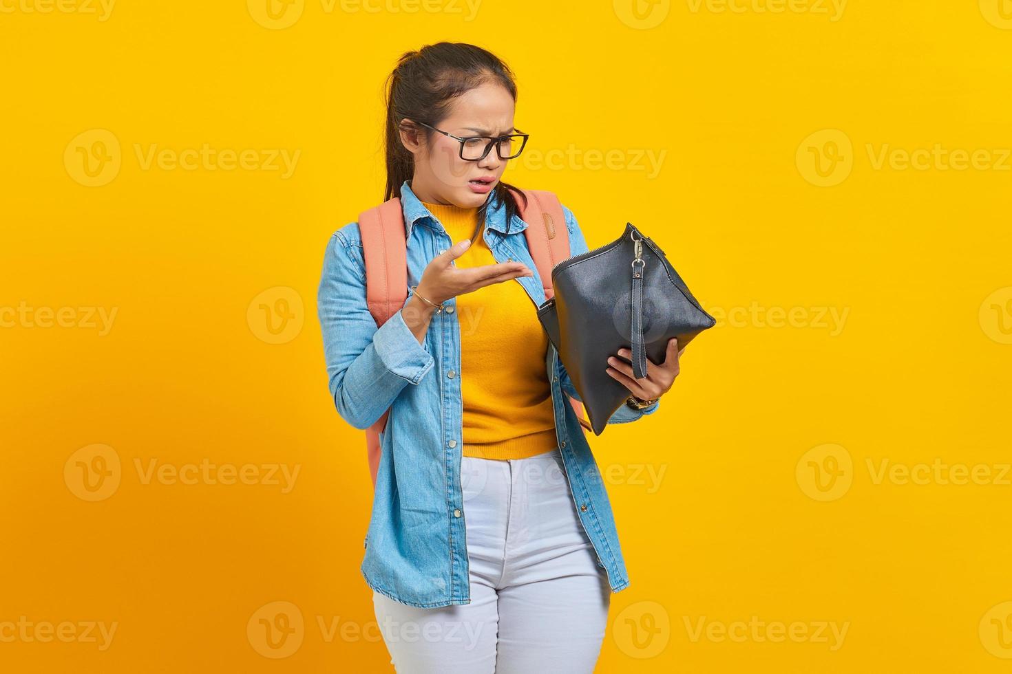 retrato de estudante surpreendida jovem mulher asiática em roupas jeans, óculos com mochila e segurando a carteira isolada em fundo amarelo. educação no conceito de faculdade universitária do ensino médio foto