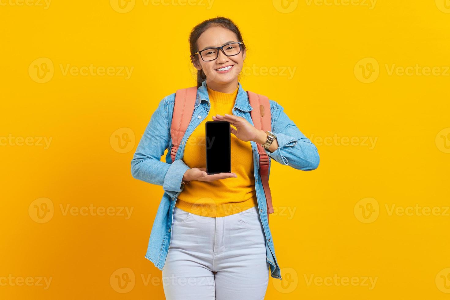 retrato de estudante asiática jovem sorridente em roupas casuais com mochila mostrando celular de tela em branco isolado em fundo amarelo. educação no conceito de universidade universitária foto