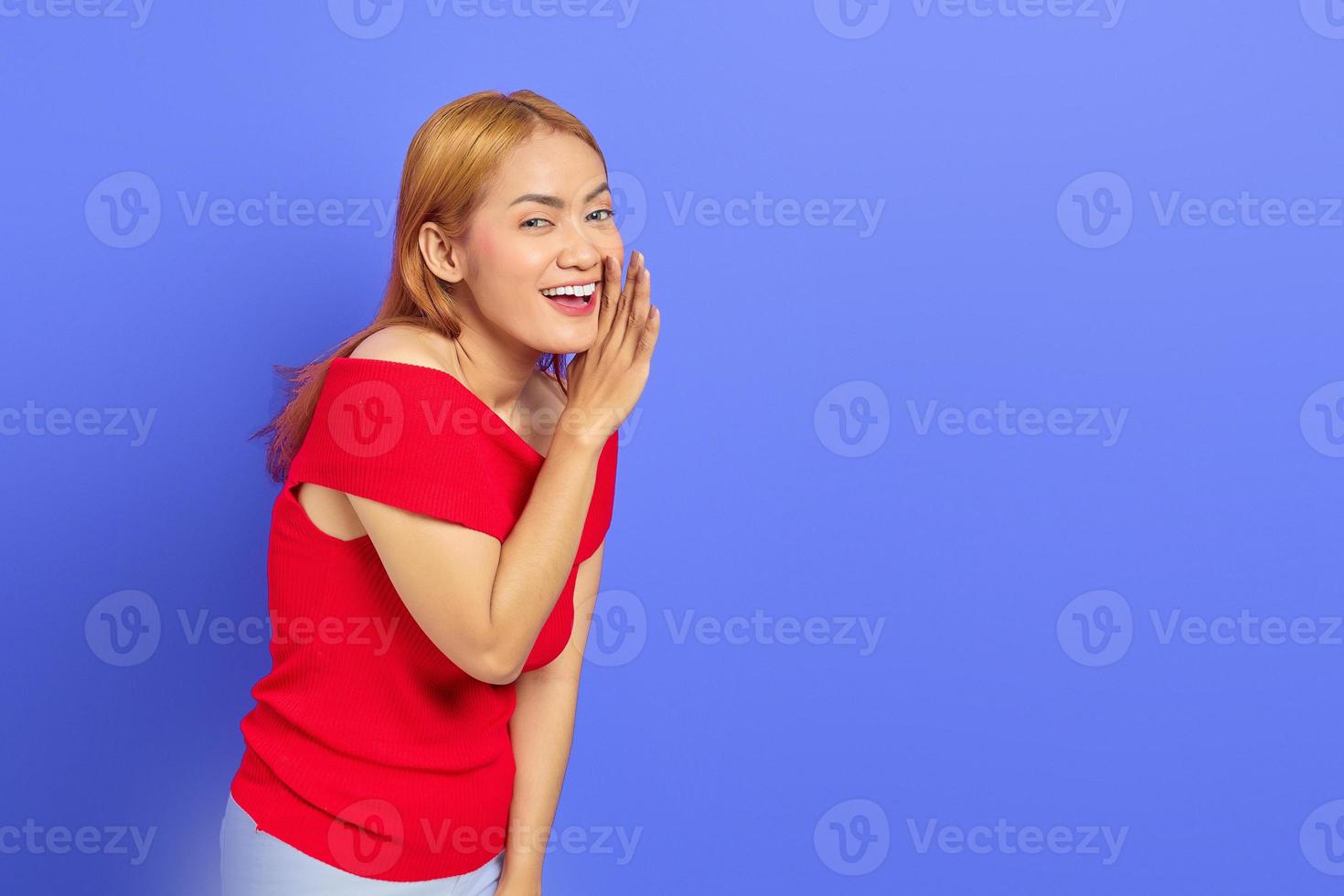 retrato de jovem mulher asiática de vestido vermelho, olhando para a câmera e sussurrando enquanto segura a mão na boca isolada sobre fundo roxo foto