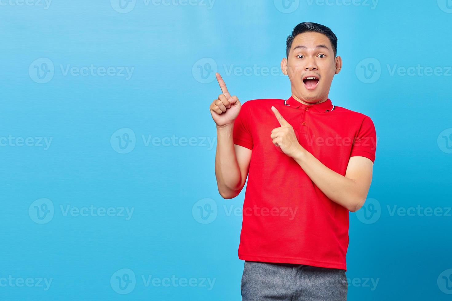 retrato de jovem asiático surpreso na camisa vermelha em pé apontando o dedo no espaço de cópia isolado no fundo azul foto