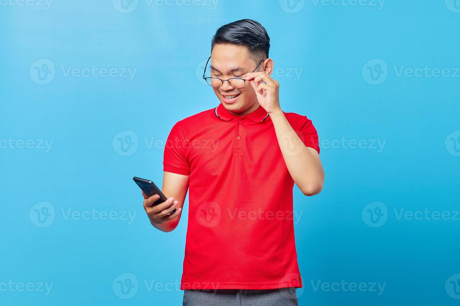 retrato de jovem asiático sorridente segurando óculos e lendo a mensagem recebida no celular isolado em fundo azul foto