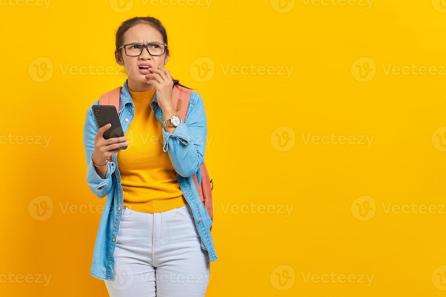 retrato de estudante asiática jovem séria em roupas casuais com mochila usando telefone celular e roendo as unhas enquanto olha de lado isolado em fundo amarelo foto