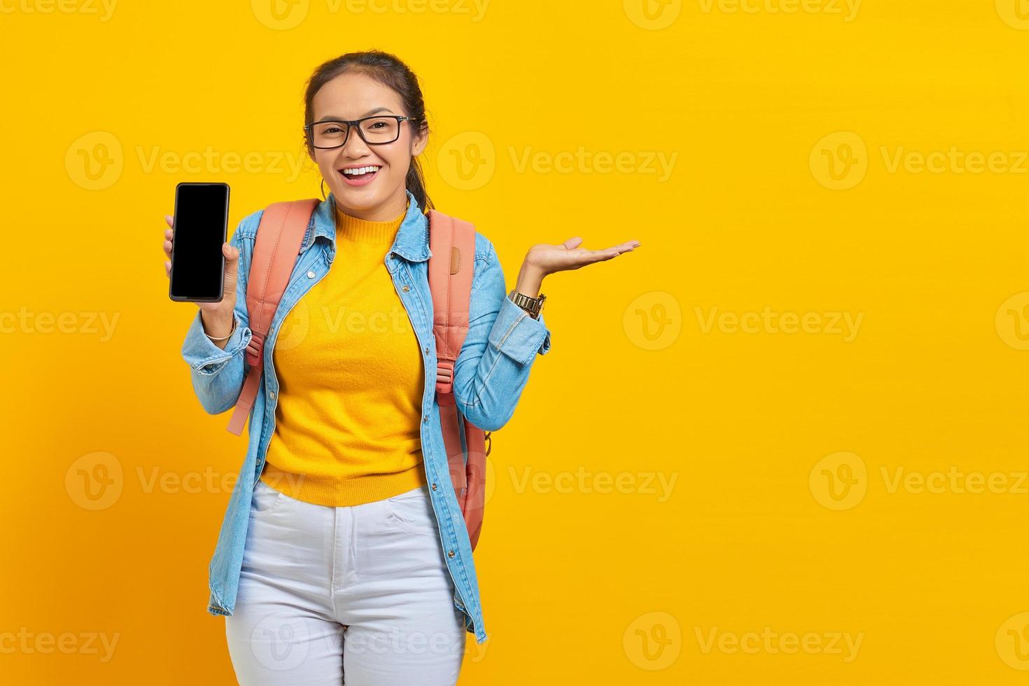 retrato de estudante asiática jovem alegre em roupas casuais com mochila segurando smartphone e apontando para copiar o espaço com as palmas das mãos isoladas em fundo amarelo foto