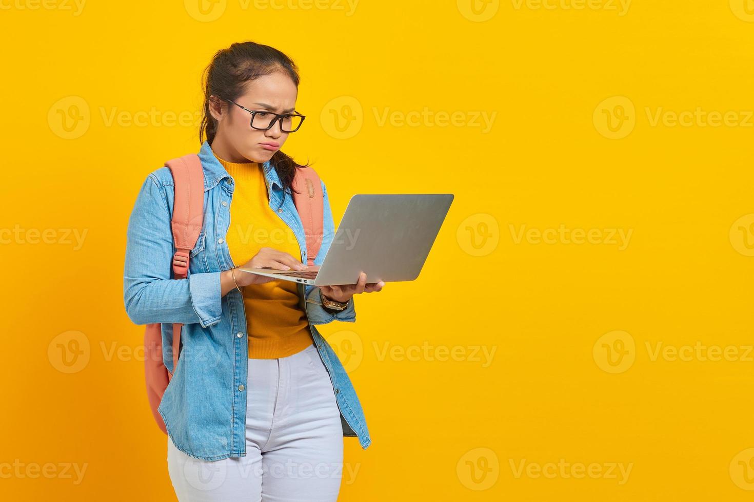 retrato de estudante asiática jovem confusa em roupas casuais com mochila olhando e-mail de entrada no laptop isolado em fundo amarelo. educação no conceito de faculdade universitária foto