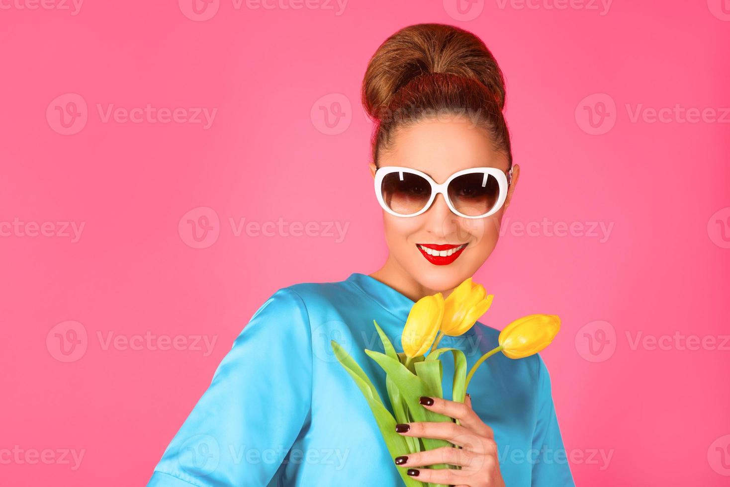 retrato de uma jovem linda de vestido de seda azul e óculos de sol brancos com tulipas amarelas no fundo rosa foto