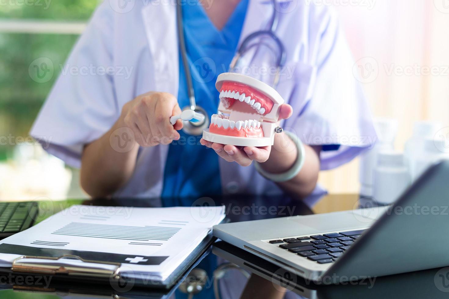 dentista concentrado sentado à mesa com amostras de mandíbula modelo de dente e trabalhando com tablet e laptop em consultório odontológico foto