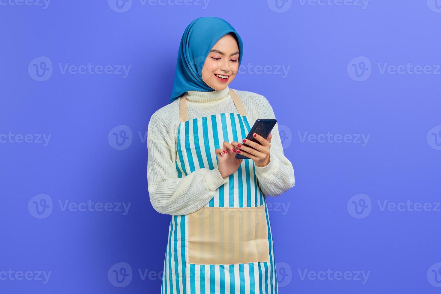 retrato de sorridente jovem muçulmana asiática em 20 anos vestindo hijab e avental digitando mensagens sms no celular isolado no fundo roxo. conceito de estilo de vida muçulmano dona de casa de pessoas foto
