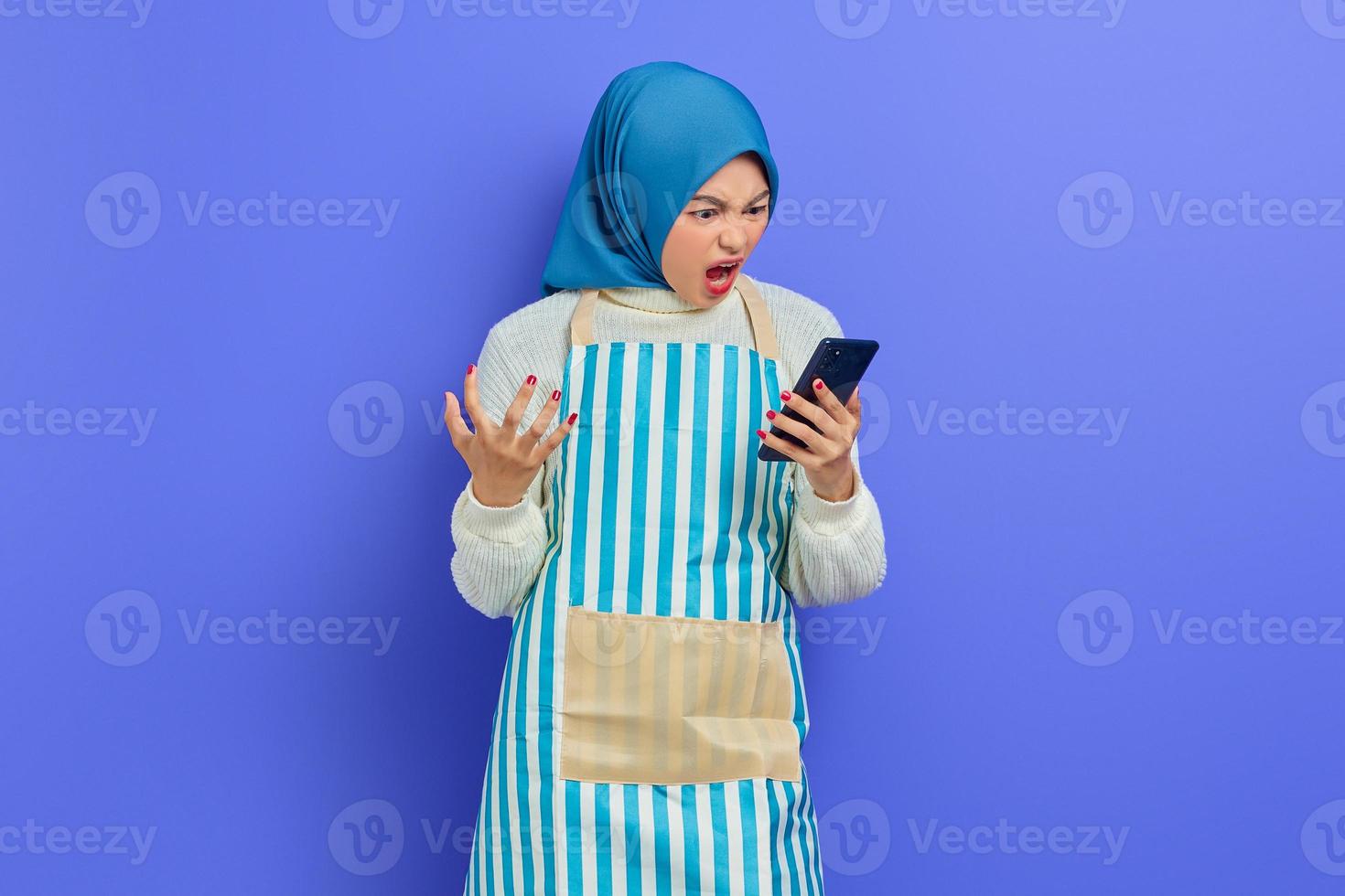 retrato de mulher muçulmana asiática jovem com raiva vestindo hijab e avental usando telefone celular enquanto gritava alto isolado sobre fundo roxo. conceito de estilo de vida muçulmano dona de casa de pessoas foto