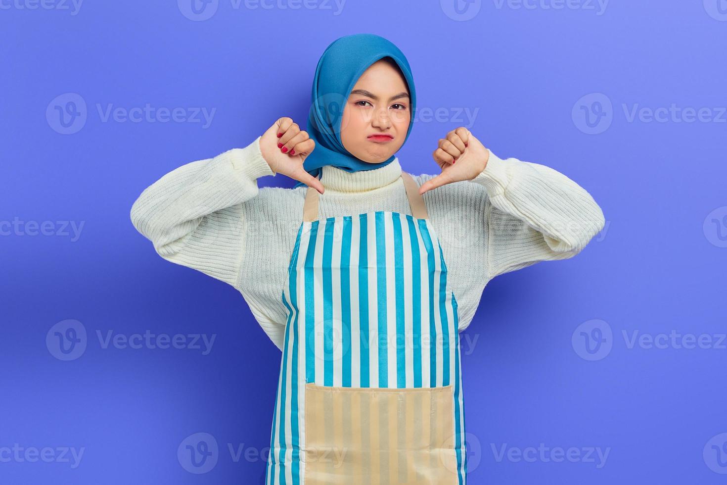 retrato de mulher jovem dona de casa descontente em hijab e avental listrado, posando mostrando o polegar para baixo enquanto olha para a câmera isolada no fundo roxo. conceito de estilo de vida muçulmano dona de casa de pessoas foto