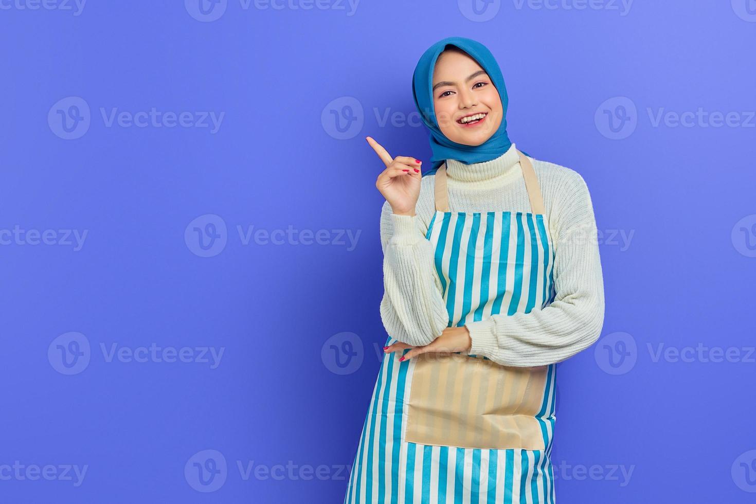 retrato de mulher jovem dona de casa sorridente em hijab e avental em pé segurando o dedo indicador com grande ideia nova, olhando a câmera no fundo roxo. conceito de estilo de vida muçulmano dona de casa de pessoas foto