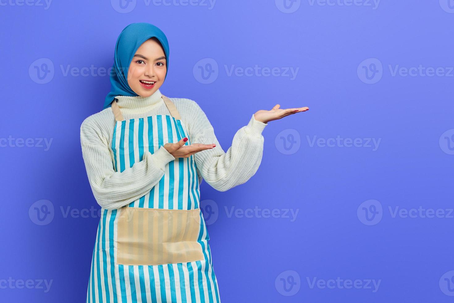 retrato de sorridente jovem muçulmana asiática em hijab e avental listrado, apontando de lado com as mãos ao fazer trabalhos domésticos isolados no fundo roxo. conceito de estilo de vida muçulmano dona de casa de pessoas foto