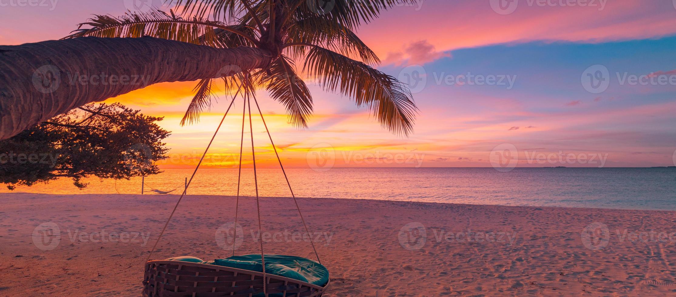 praia do sol tropical como panorama de paisagem de verão com balanço de praia ou rede. céu colorido, folhas de palmeira de areia e bandeira de praia de mar calmo. cena de praia romântica de casal foto