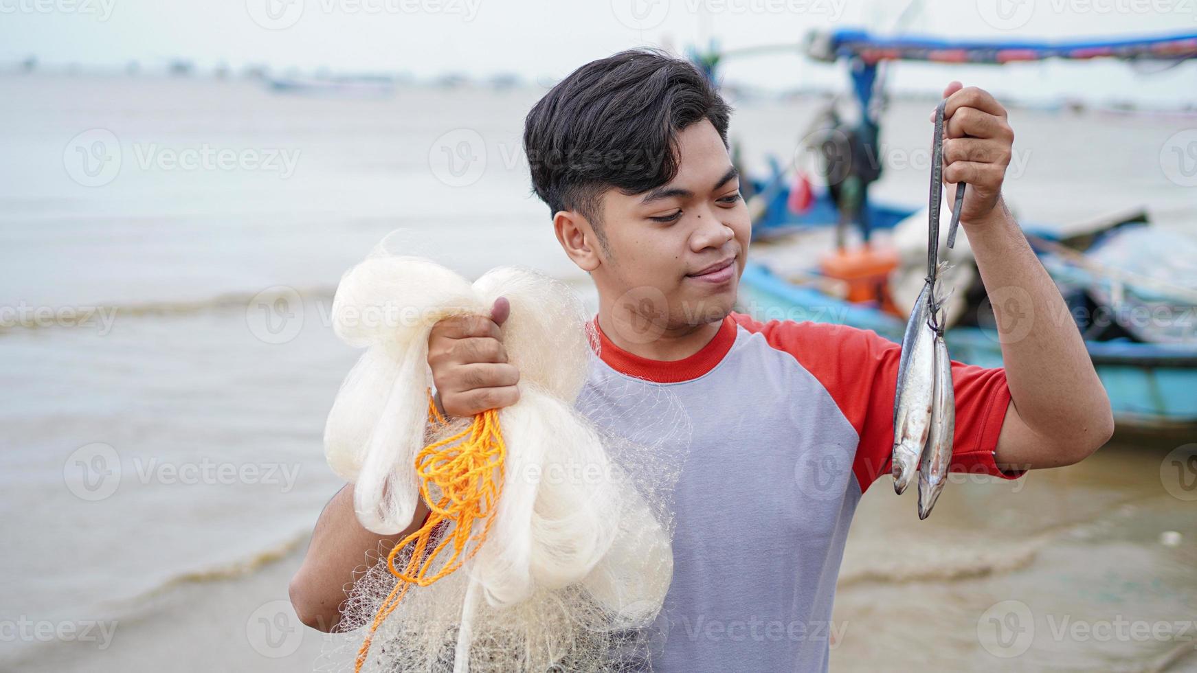 feliz jovem pescador na praia segurando seu peixe e mostra na frente de seu barco foto
