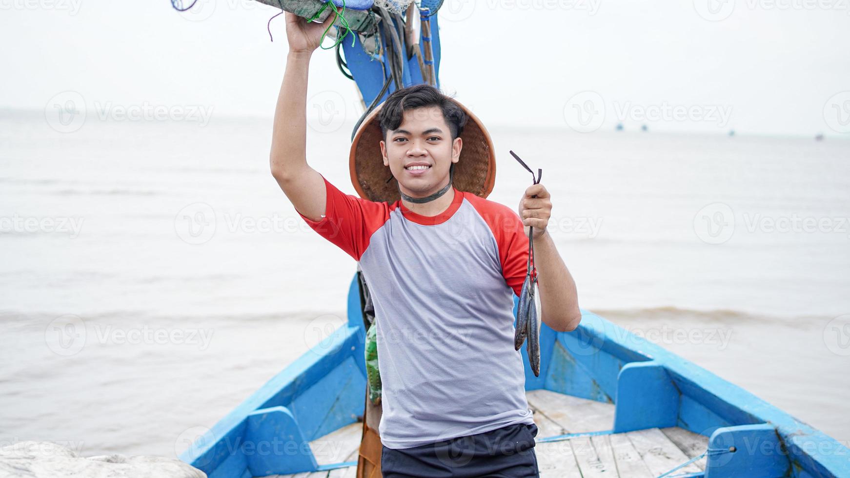 feliz jovem pescador na praia segurando seu peixe e mostra na frente de seu barco foto
