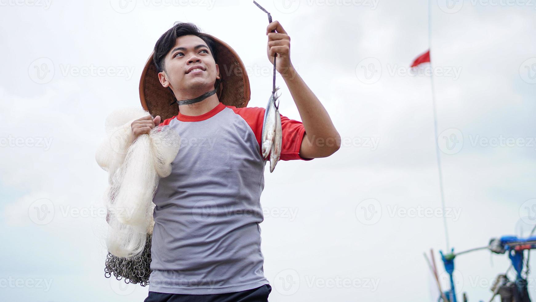 feliz jovem pescador na praia segurando seu peixe e mostra na frente de seu barco foto