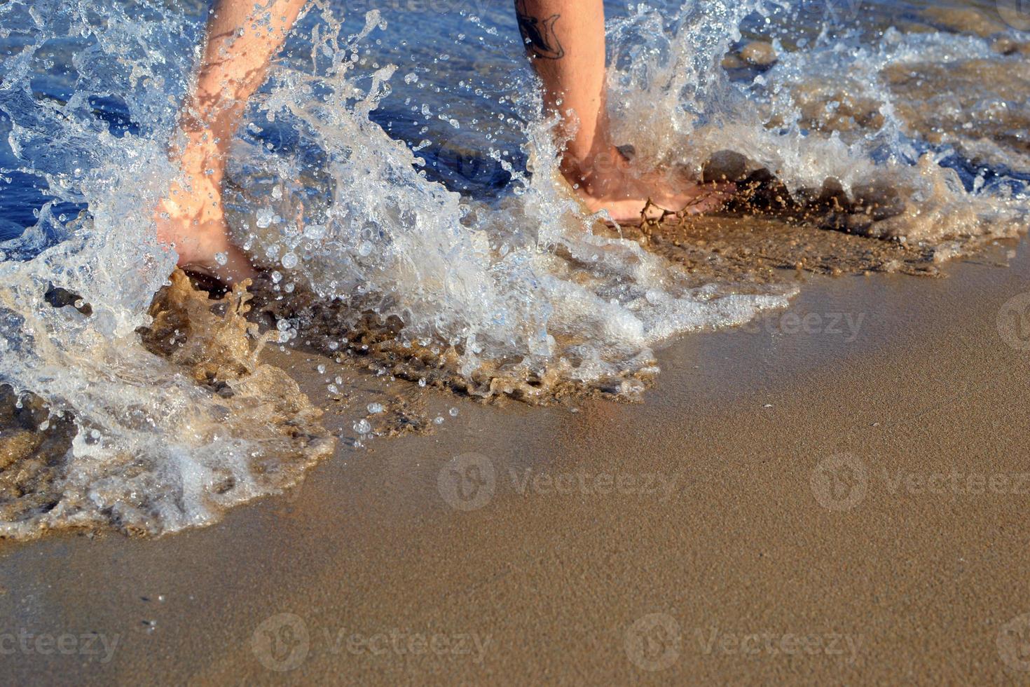 a onda quebra nas pernas da garota. pernas de uma linda garota correndo para o oceano em uma praia arenosa. pés femininos acenando na onda do mar. férias em praias paradisíacas.espuma oceânica envolve a menina foto