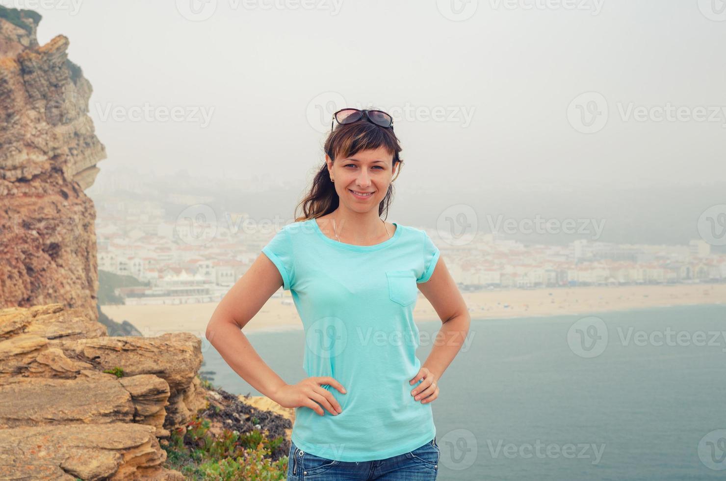 jovem viajante bonita olhando para a câmera posando e sorrindo para falésias e rochas de pedra da cidade de nazaré foto