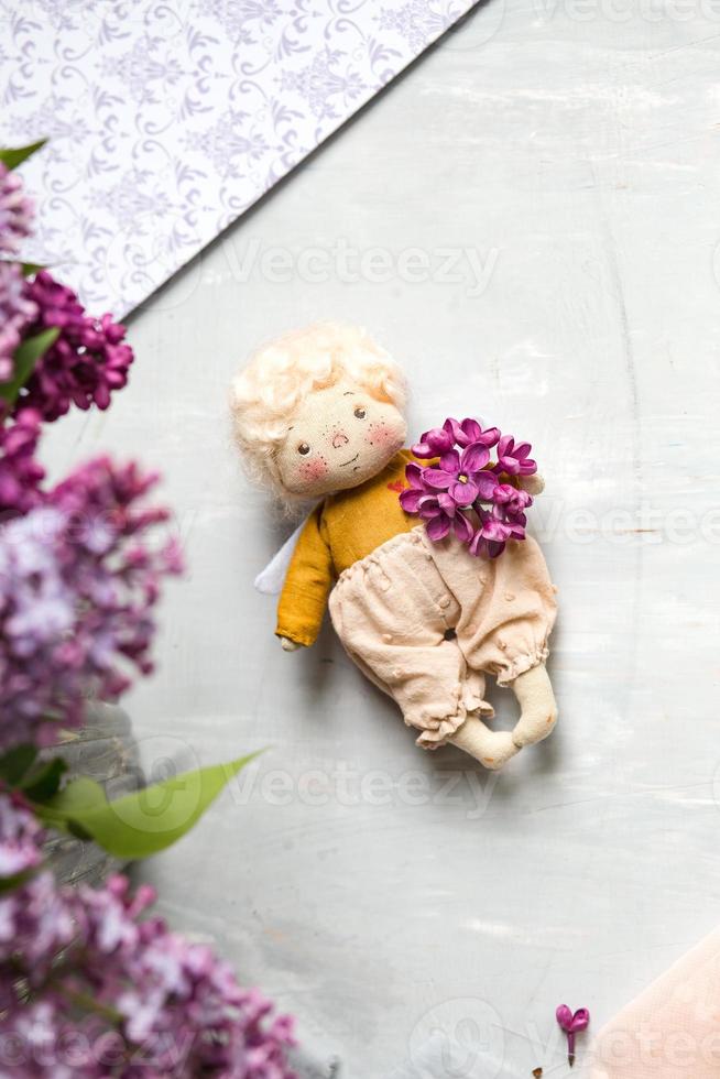 anjinho de cabelos dourados nas flores lilás azul, rosa, roxo, violeta. brinquedo artesanal em cores lilás violetas. cartão de saudação. foto