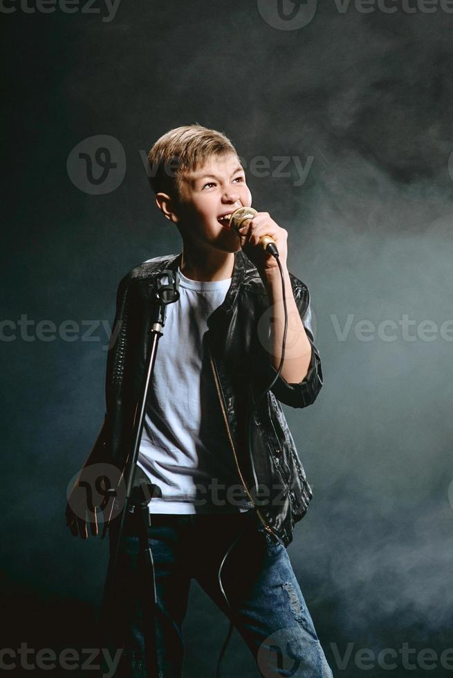 retrato de adolescente caucasiano em camiseta branca, jeans azul e jaqueta de couro com microfone cantando em fundo escuro. conceito de hobby e glória foto