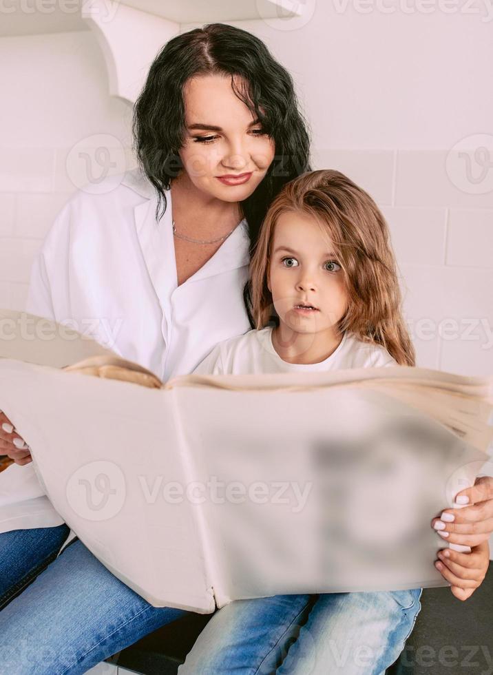 linda mulher morena elegante com adorável filha lendo notícias de última hora no jornal de papel. família, jornal, notícias, choque, amor, conceito de apoio foto