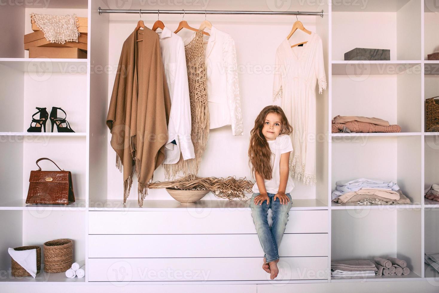 menina engraçada em t-shirt branca e jeans, sentado no guarda-roupa, sonhando em ser adulto e elegante. idade, infância, moda, conceito de estilo foto