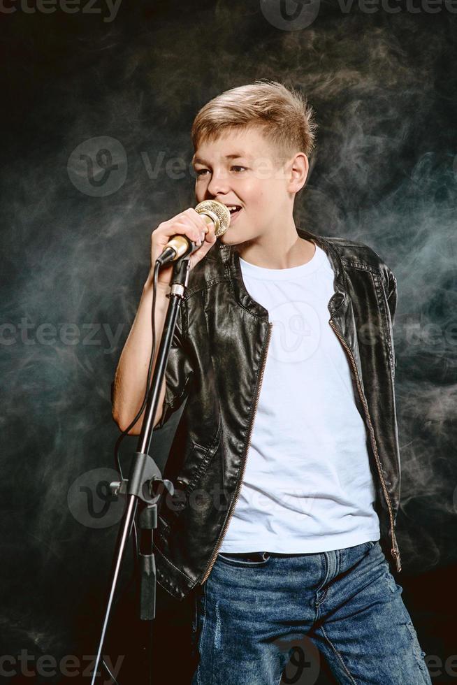 retrato de adolescente caucasiano em camiseta branca, jeans azul e jaqueta de couro com microfone cantando em fundo escuro. conceito de hobby e glória foto