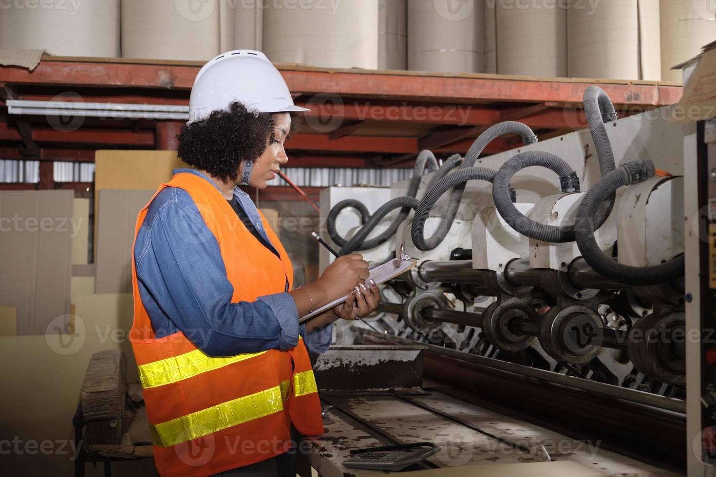 uma trabalhadora industrial afro-americana com um capacete verifica a máquina e escreve peças de motor inspecionadas. profissional trabalha em uma fábrica de fabricação de papel, mantém equipamentos de produção. foto