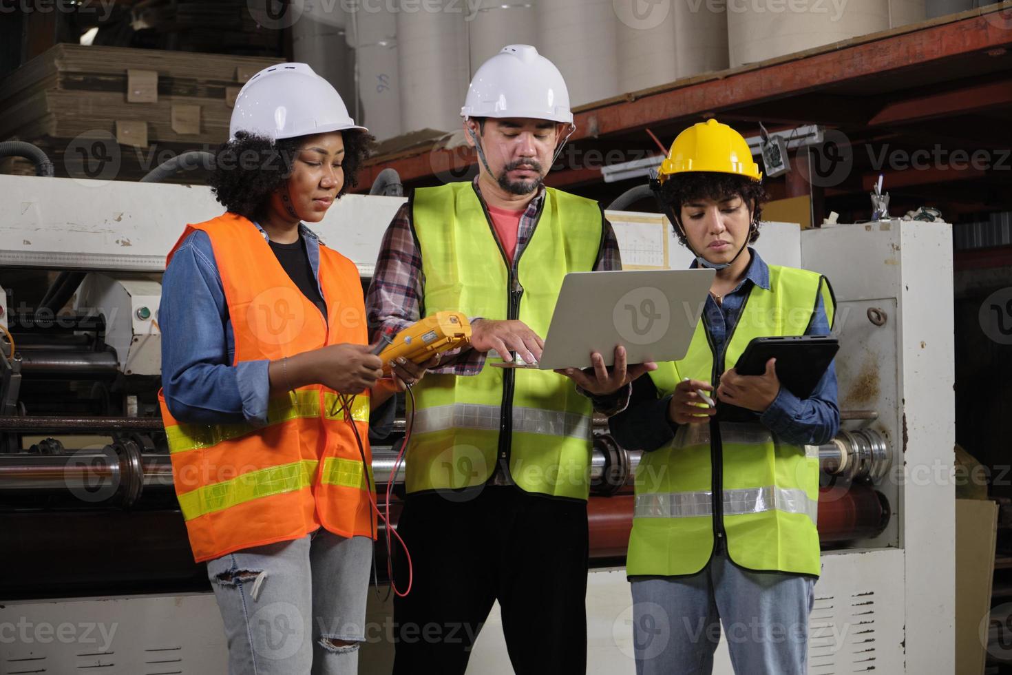 trabalhadores de uniformes de segurança e engenheiros industriais com capacete de segurança usam computador portátil para verificar e controlar máquinas. três profissionais trabalham na fábrica de fabricação de papel, mantêm equipamentos de produção. foto