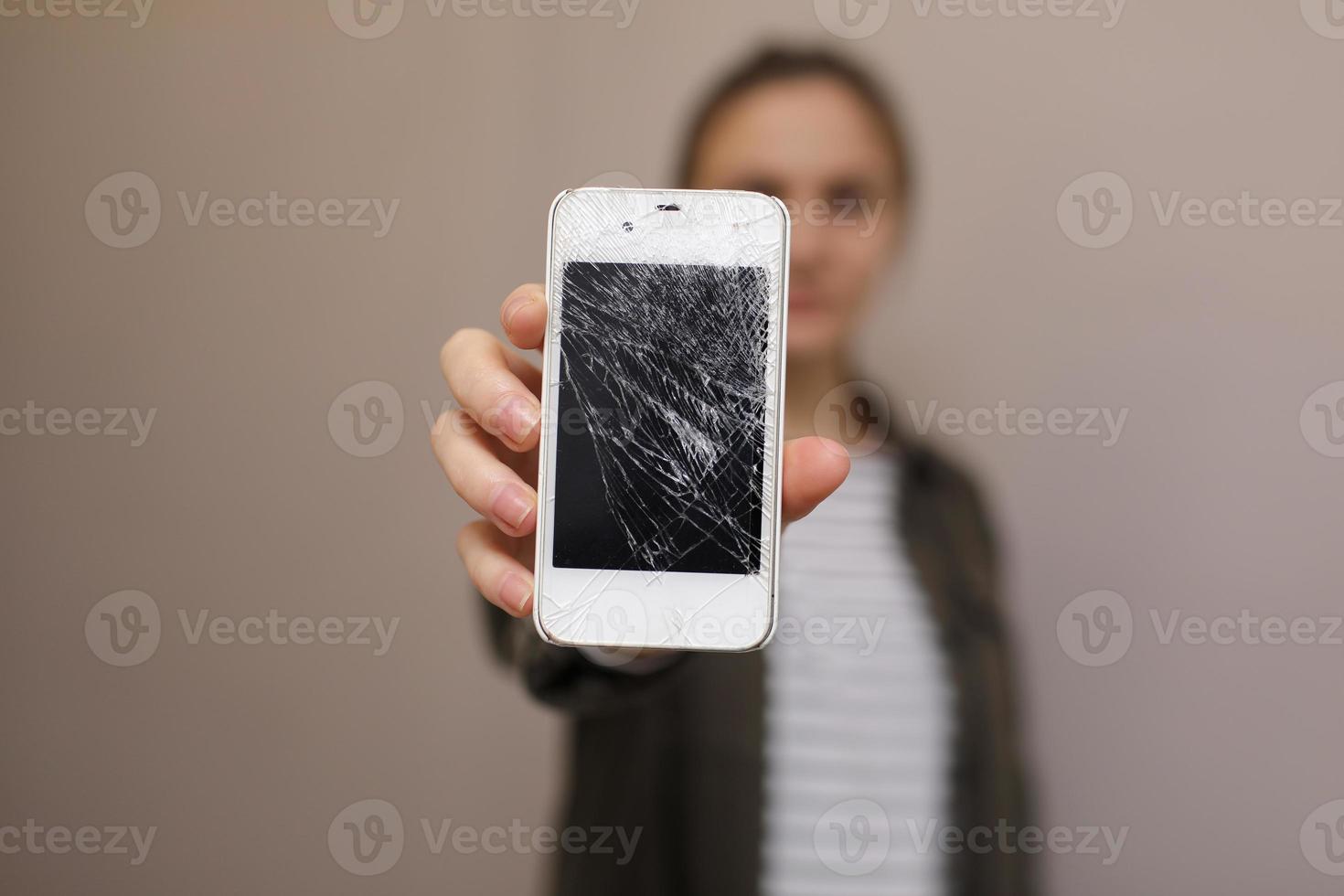 jovem frustrada segurando o celular com o vidro da tela quebrada. a tela do telefone precisa ser consertada. isolado em fundo cinza. foco seletivo no smartphone foto