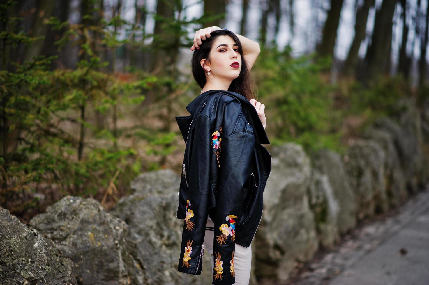 menina cigana morena posou contra pedras no parque. desgaste do modelo na jaqueta de couro e t-shirt com ornamento, calças. foto