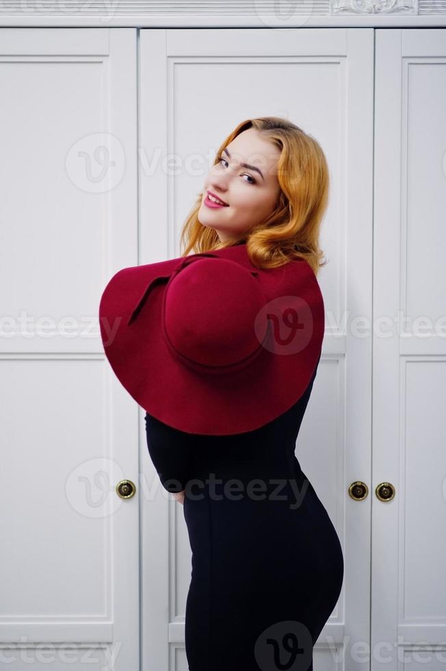 retrato de moda garota ruiva no chapéu vermelho e vestido preto com maquiagem brilhante posou perto do armário de madeira vintage branco. filtros de instagram de estilo de foto em tons.