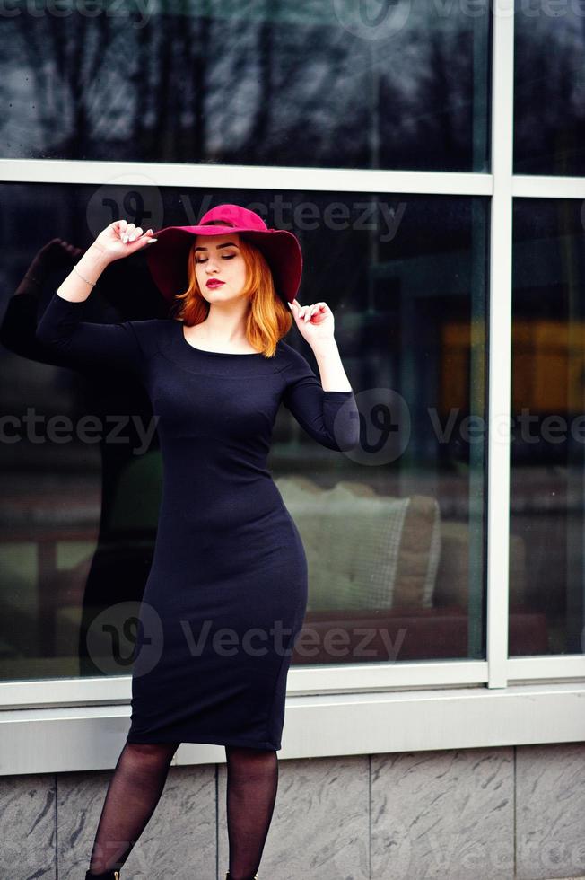 retrato de moda garota ruiva no chapéu vermelho e vestido preto com maquiagem brilhante posou contra a grande janela. filtros de instagram de estilo de foto em tons.