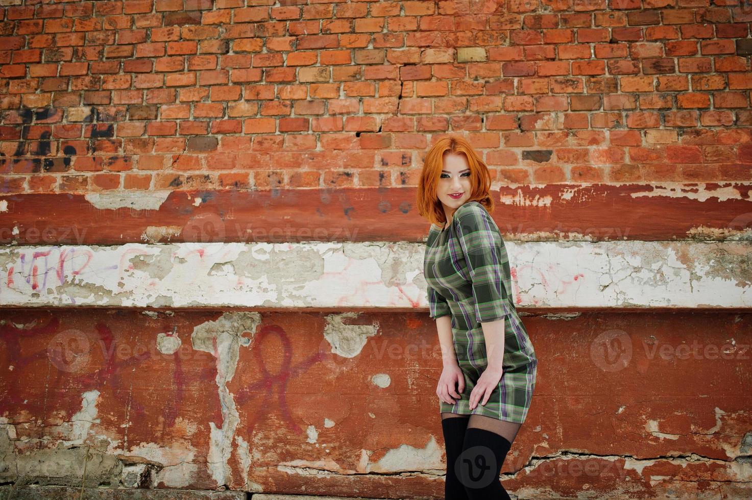 um retrato ao ar livre de uma jovem bonita com cabelo vermelho, vestido xadrez em pé sobre o fundo da parede de tijolo em dia de inverno. foto