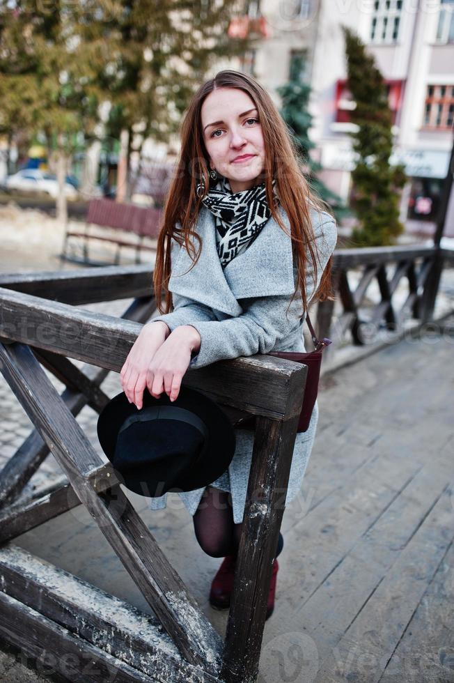 garota jovem modelo de casaco cinza e chapéu preto com bolsa de couro nos ombros posou contra vigas de madeira na rua da cidade. foto