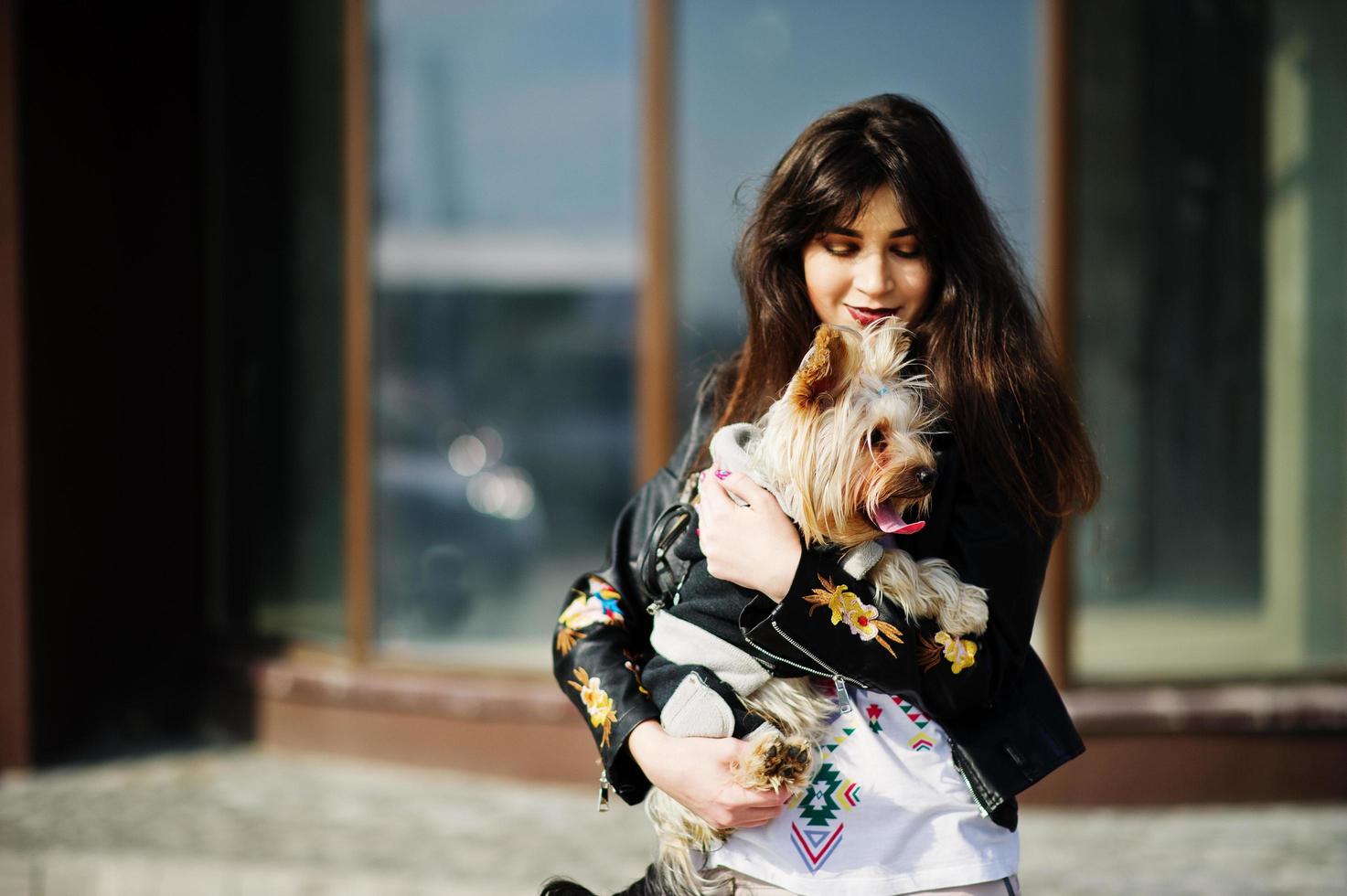 menina cigana morena com cachorro yorkshire terrier posou contra casa de janelas grandes. desgaste do modelo na jaqueta de couro e t-shirt com ornamento, calças. foto