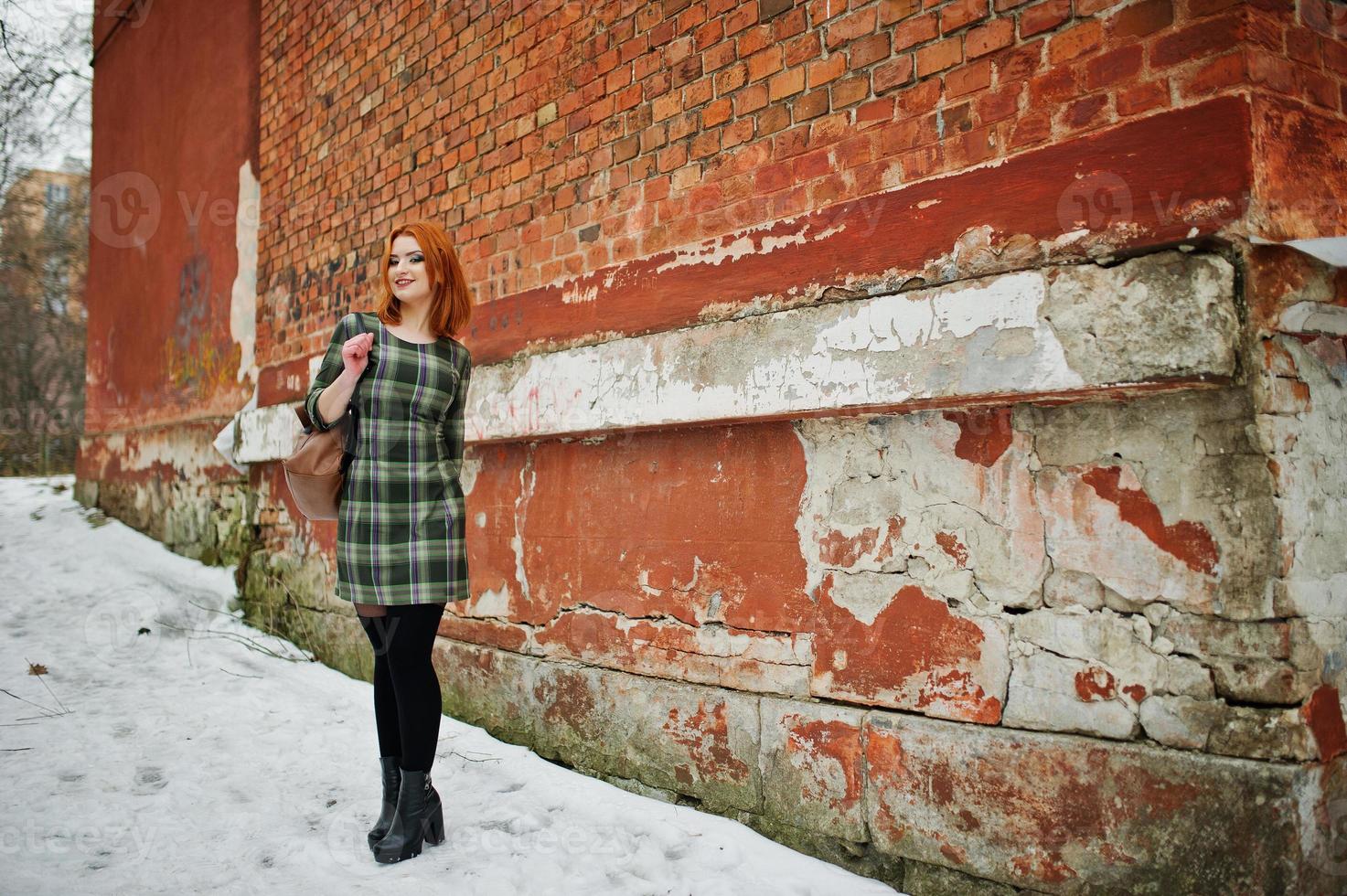 um retrato ao ar livre de uma jovem bonita com cabelo vermelho, vestido xadrez com mochilas femininas em pé no fundo da parede de tijolo em dia de inverno. foto