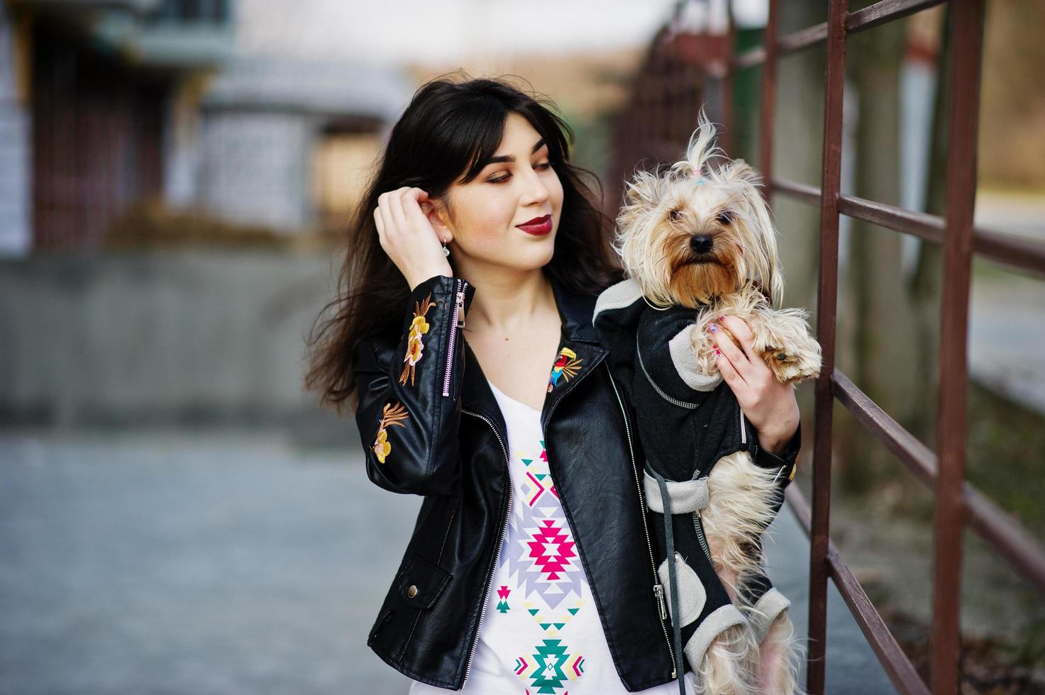 menina cigana morena com cachorro yorkshire terrier posou contra grades de aço. desgaste do modelo na jaqueta de couro e t-shirt com ornamento, calças. foto