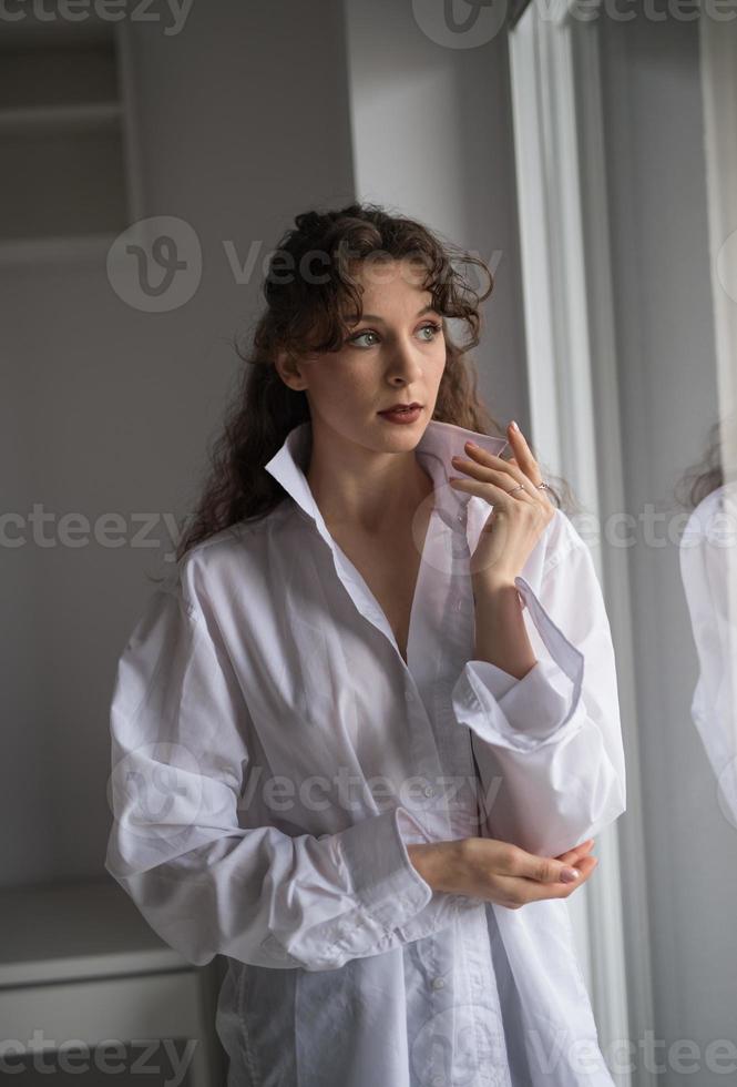 morena sexy atraente na camisa branca posando provocativamente, perto de uma janela, studio shot. retrato de mulher sensual com cabelo comprido, na cena clássica de boudoir, olhou pela janela foto