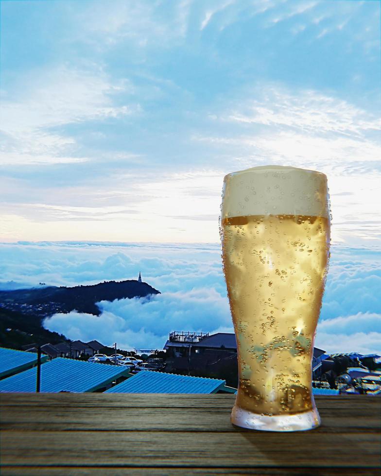 copo de cerveja transparente com cerveja bem gelada e espuma na boca do copo e gotas de água aderem às bordas. prancha ou superfície de madeira e com mar de neblina ao sol da manhã. renderização em 3D foto