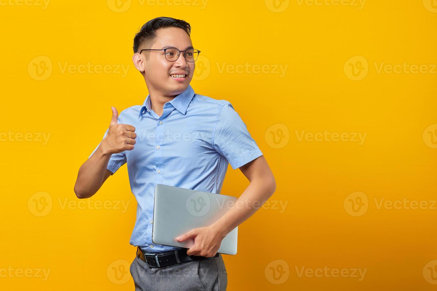 retrato de homem asiático bonito sorridente em óculos segurando laptop e fazendo o polegar no gesto, aprovar coisa boa isolada em fundo amarelo. conceito de empresário e empresário foto