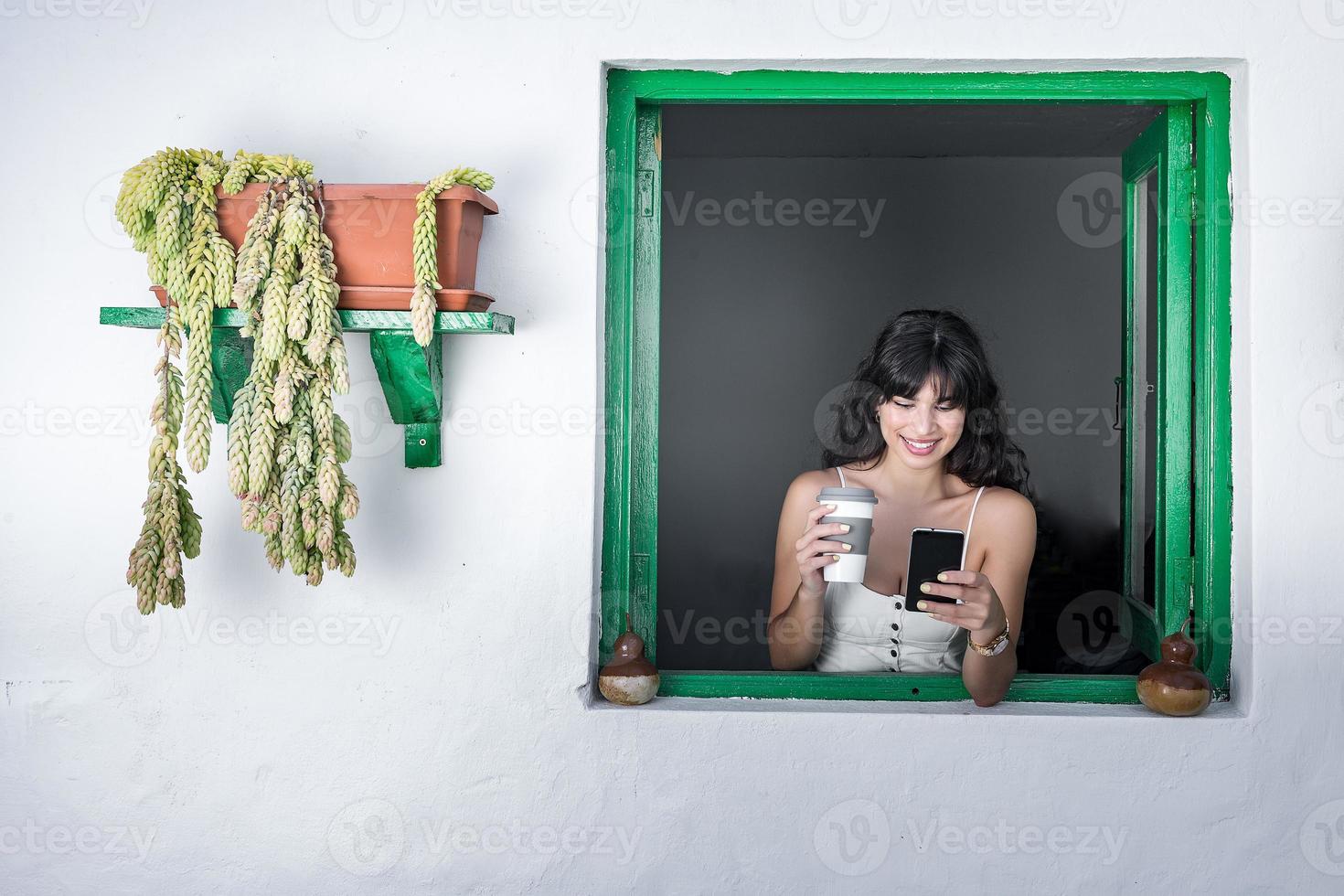 alegre mulher étnica com bebida para ir usando smartphone na janela foto