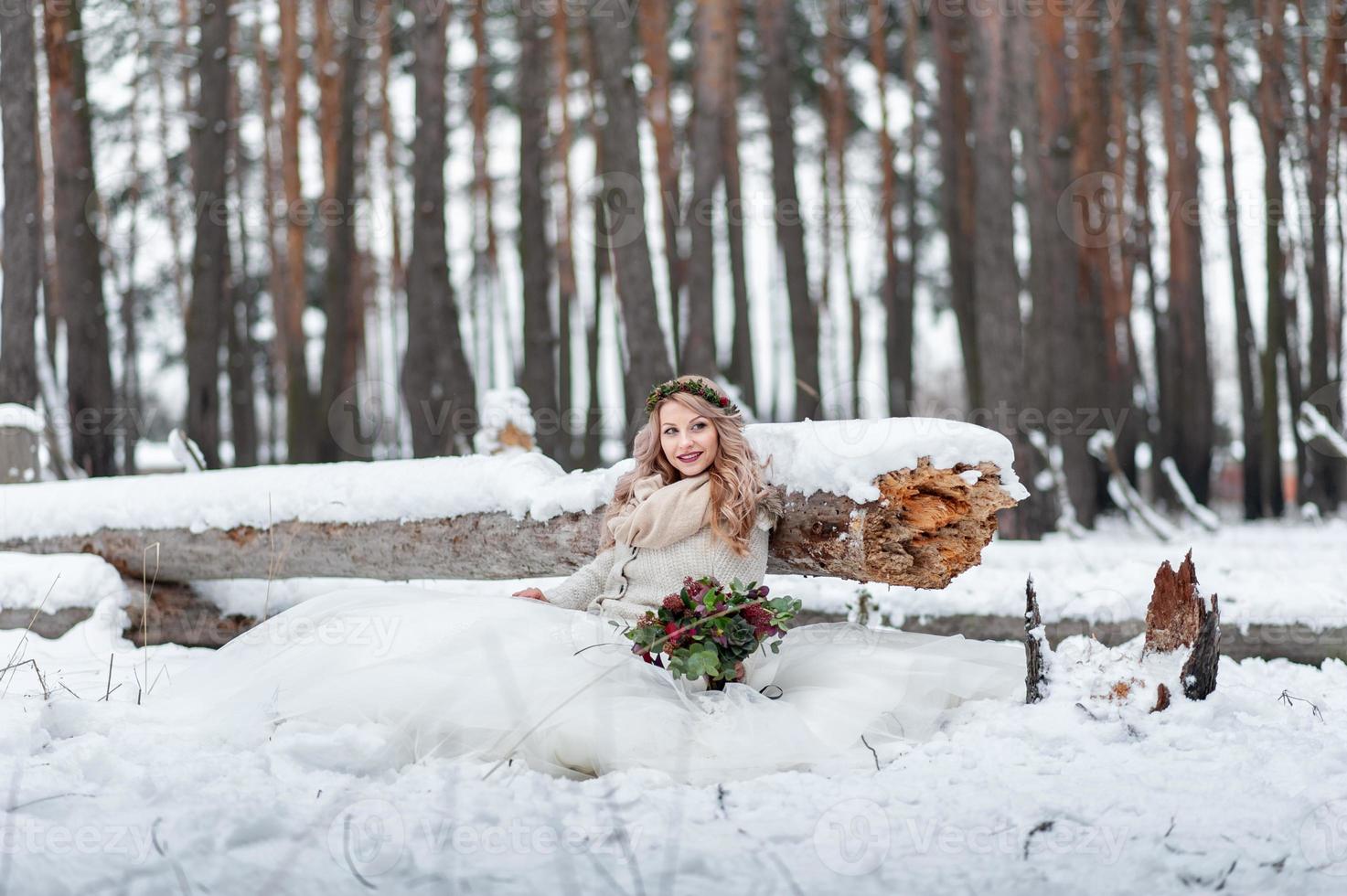 noiva bonita de aparência eslava com uma coroa de flores segura um buquê, senta-se ao lado do tronco na floresta de neve. cerimônia de casamento de inverno. foto