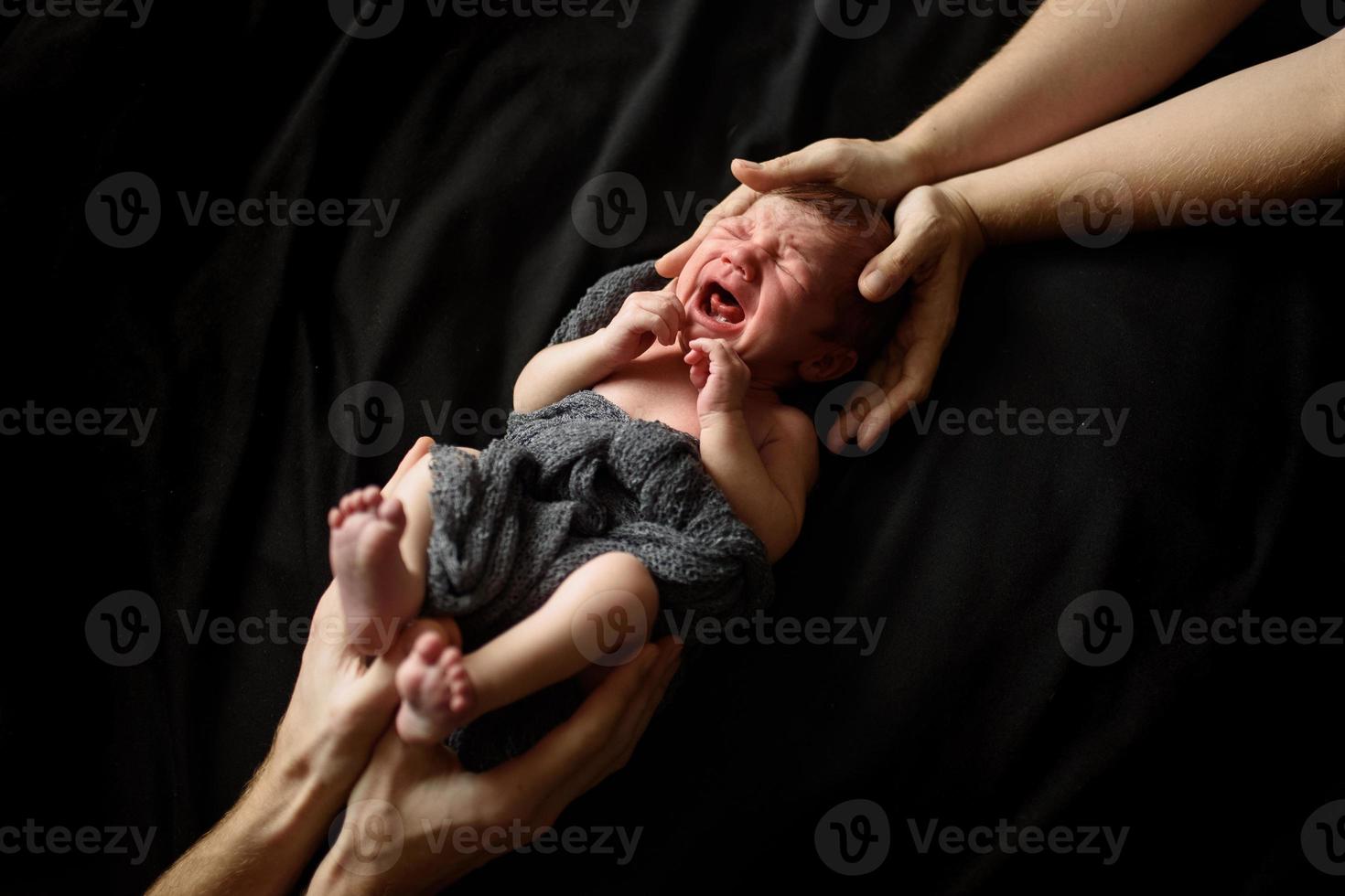 menino recém-nascido em um fundo preto. superior e inferior do menino apoiam as mãos dos pais. o bebê está chorando. foto