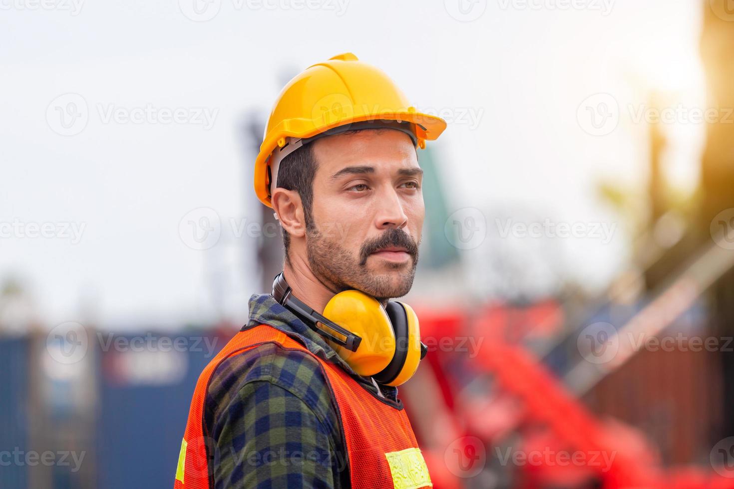 retrato de engenheiro homem de capacete e colete de segurança, trabalhador com fones de ouvido protetores no canteiro de obras foto