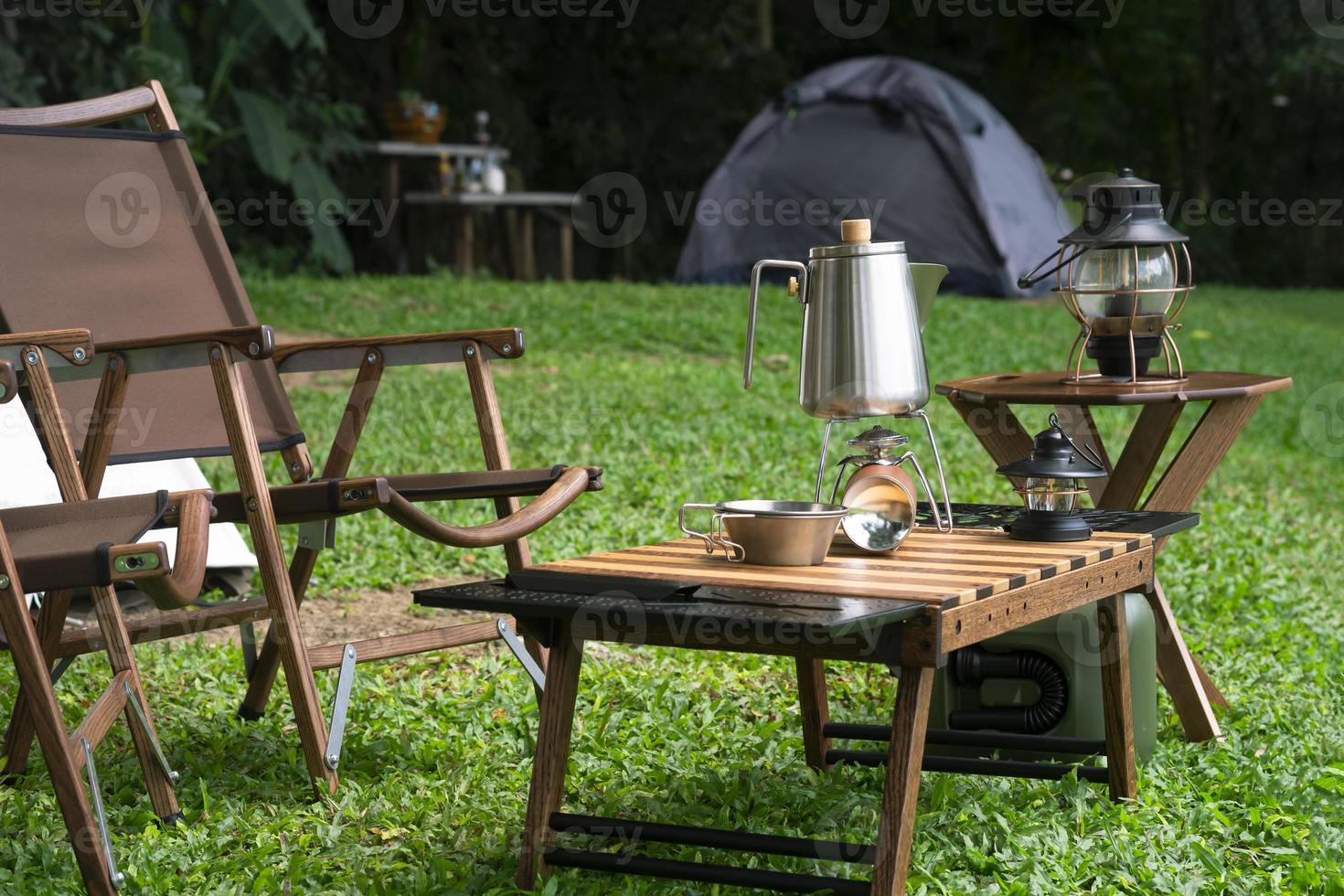 chaleira de aço inoxidável, fogão a gás portátil, tigela e lanternas vintage com mesa ao ar livre em gramado verde na área de camping foto