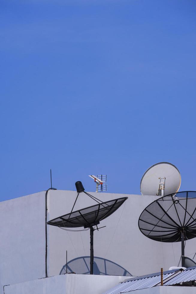 3 antenas parabólicas com antena de tv em cima do antigo prédio cinza contra o fundo azul do céu claro em quadro vertical foto