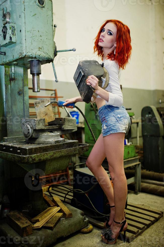 garota ruiva usa shorts jeans curtos e blusa branca com máscara de solda nas mãos posadas na máquina industrial na fábrica. foto