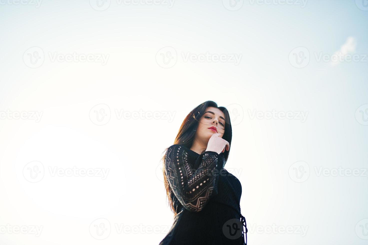 menina morena retrato com lábios vermelhos, vestindo um vestido preto posou contra o céu azul em dia ensolarado. foto