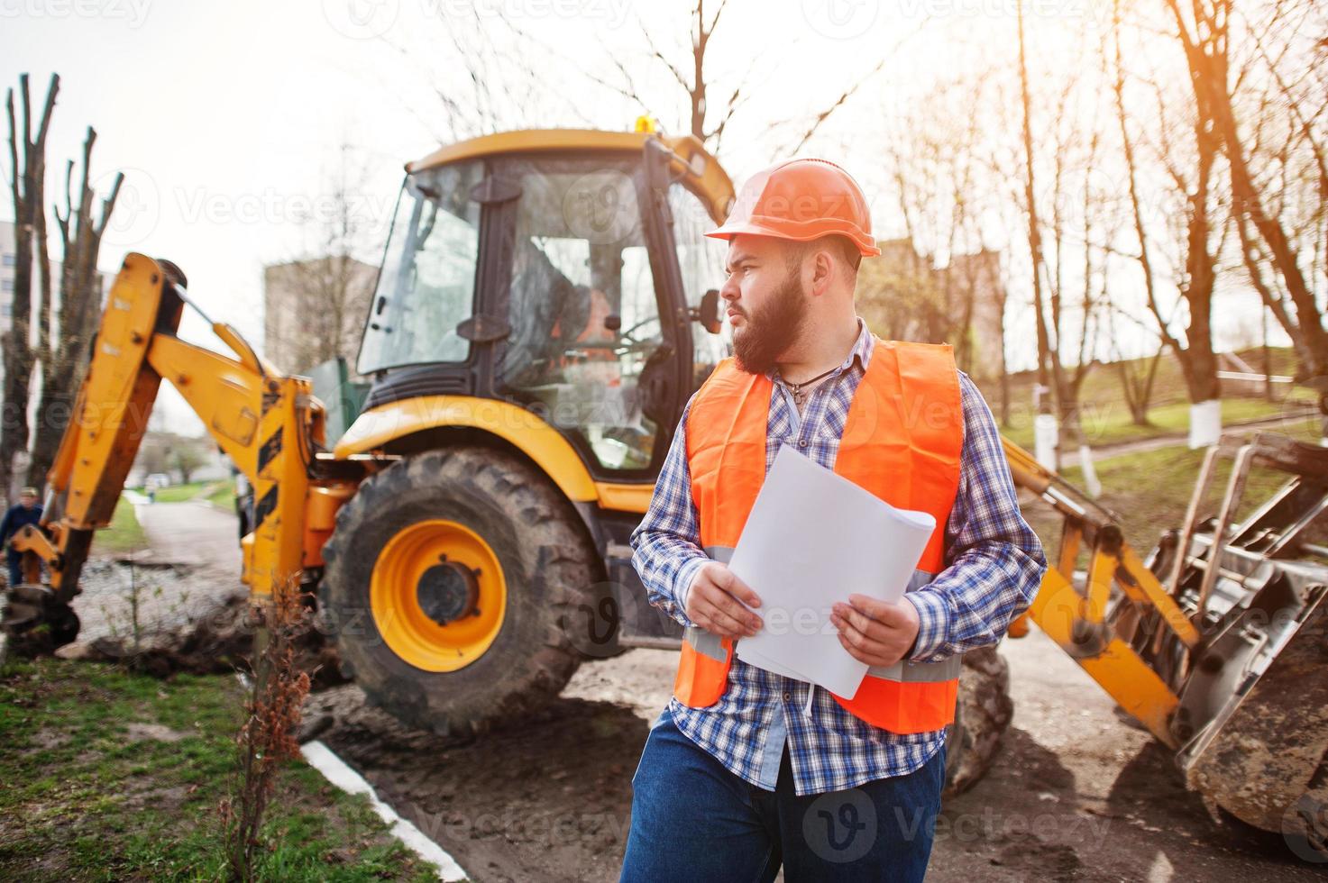 homem trabalhador barba brutal terno trabalhador da construção civil no capacete laranja de segurança contra traktor com papel de plano nas mãos. foto